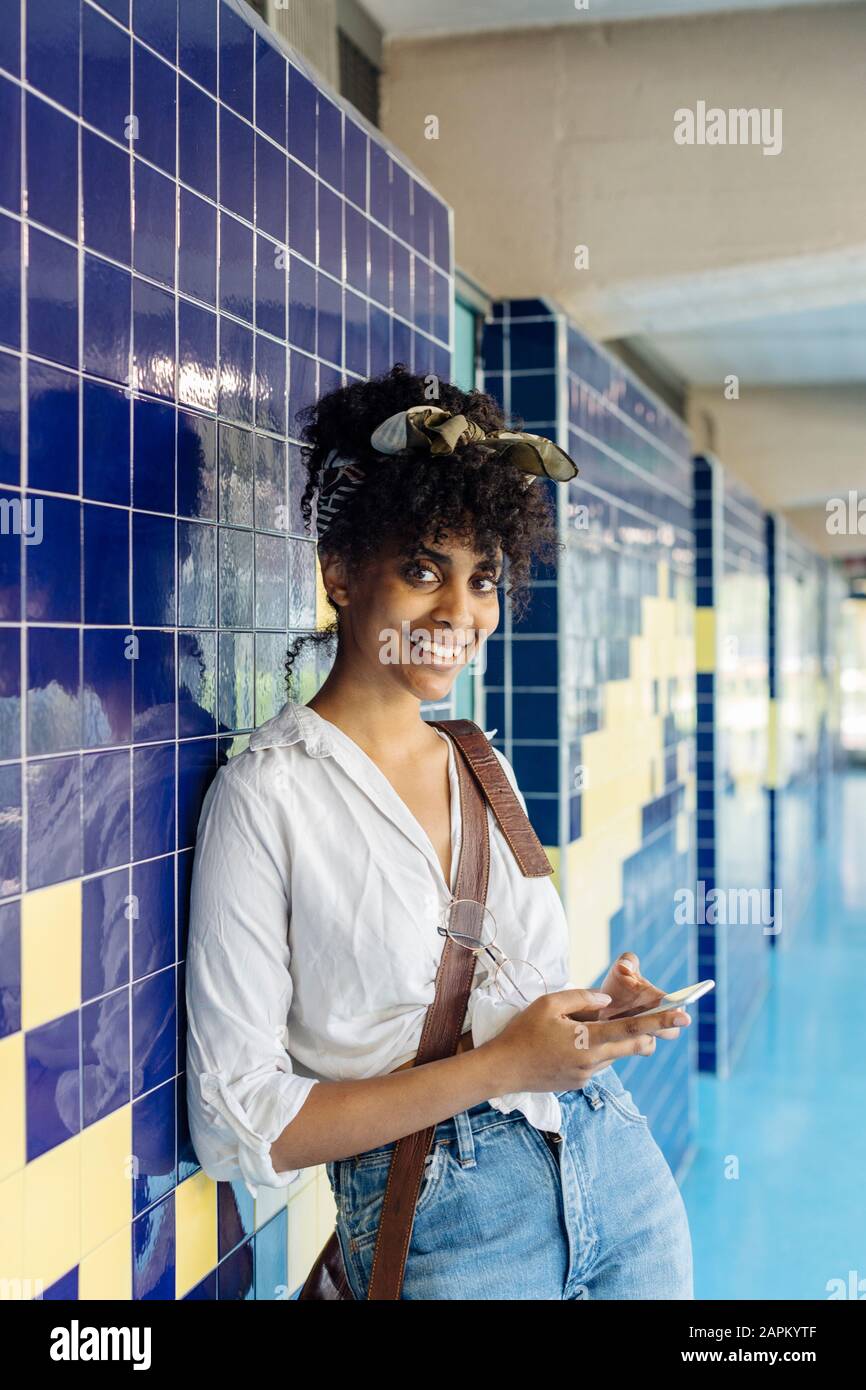 Portrait of smiling young woman with smartphone leaning against tiled wall Stock Photo