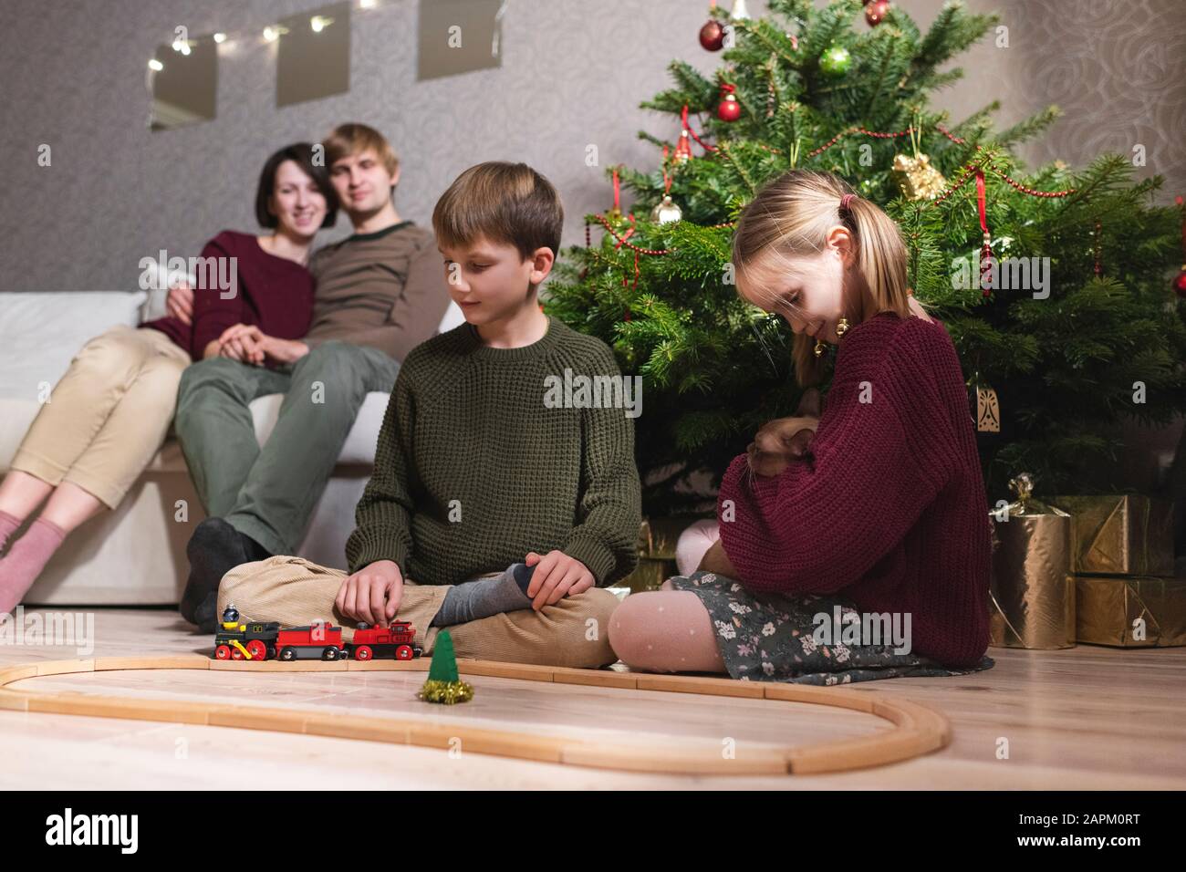 Boy and girl with catt, playing with toy train on wooden railway near Christmas tree and parents sitting on the sofa behind their children Stock Photo