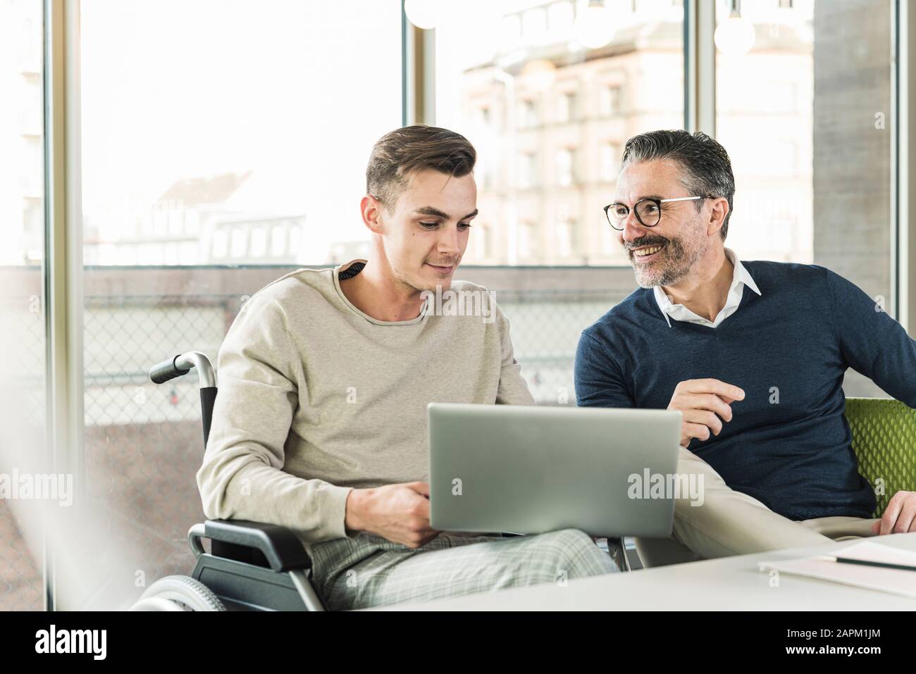Mature businessman and young man in wheelchair using laptop in office Stock Photo