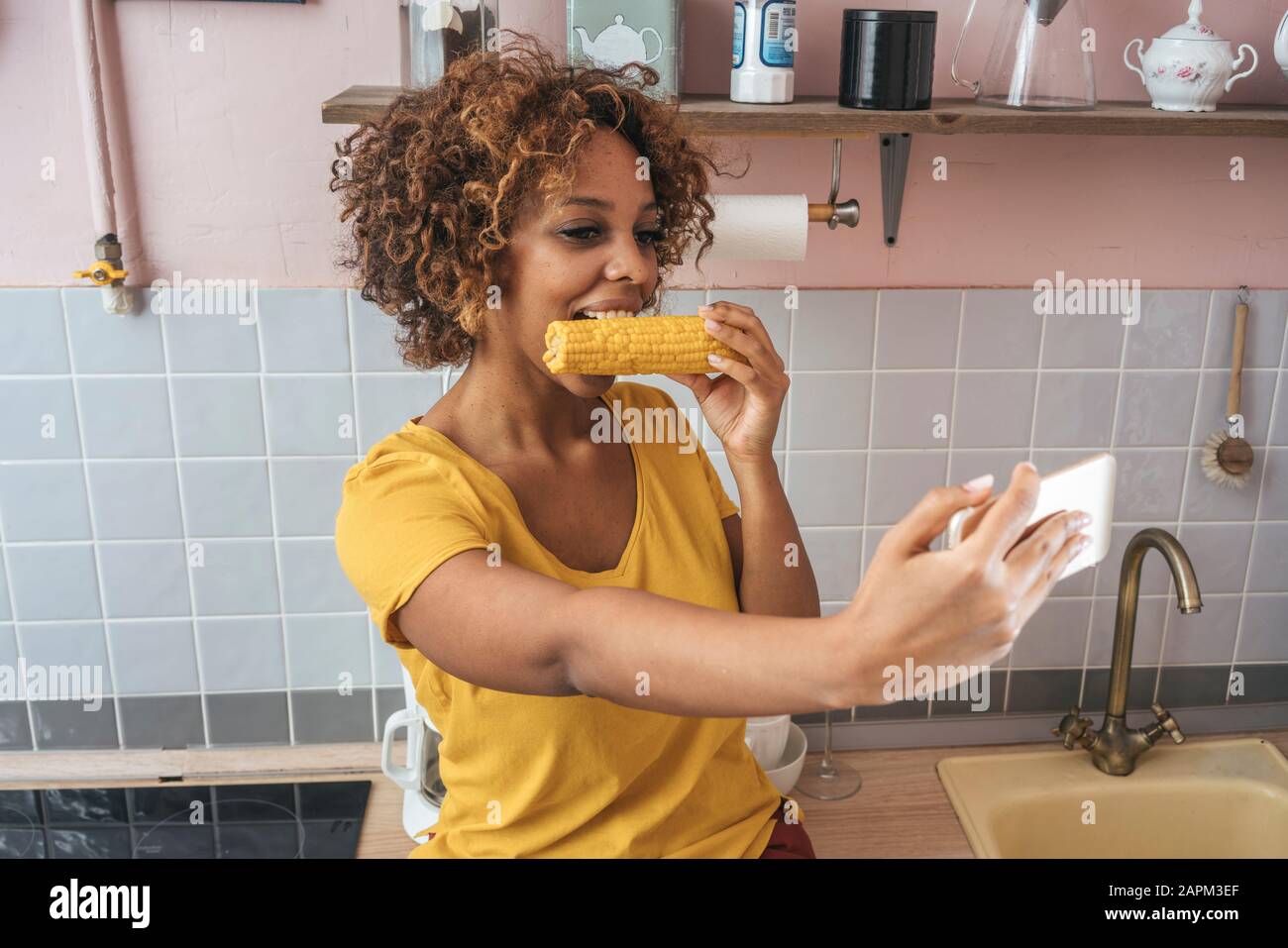 Young woman eating a corncob and taking a selfie in kitchen Stock Photo