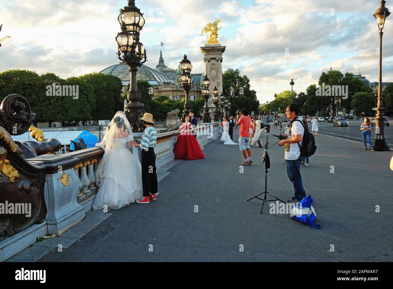 Brides, photographers, an ornate bridge, wedding dresses white or red with gold statues and art nouveau lamps, Pont Alexandre III, Paris; France Stock Photo