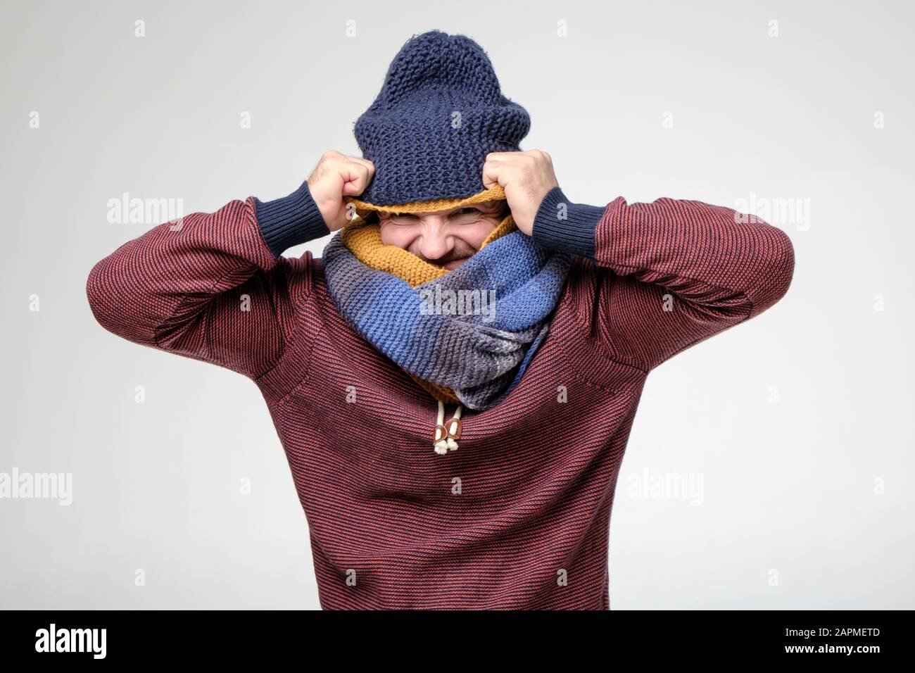 Frozen man, with a strange grimace on face, is trying to warm himself by putting on several scarves and hats. Funny fashion. Stock Photo