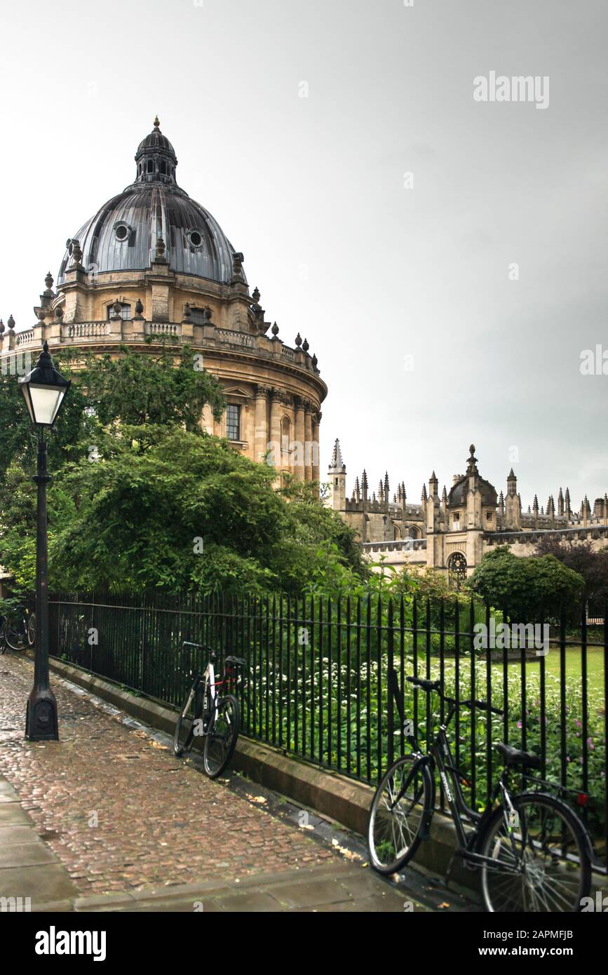 Radcliffe Camera and All Souls College on a rainy day. Bikes rest against a spiked metal railing enclosing a green lawn. Street lamp and cobbled path. Stock Photo