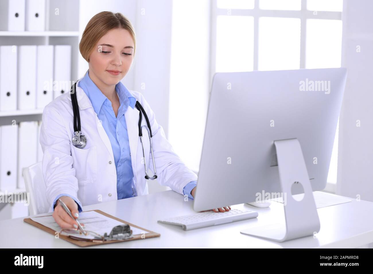 Young woman doctor at work in hospital looking at desktop pc monitor. Physician controls medication history records and exam results. Medicine and Stock Photo