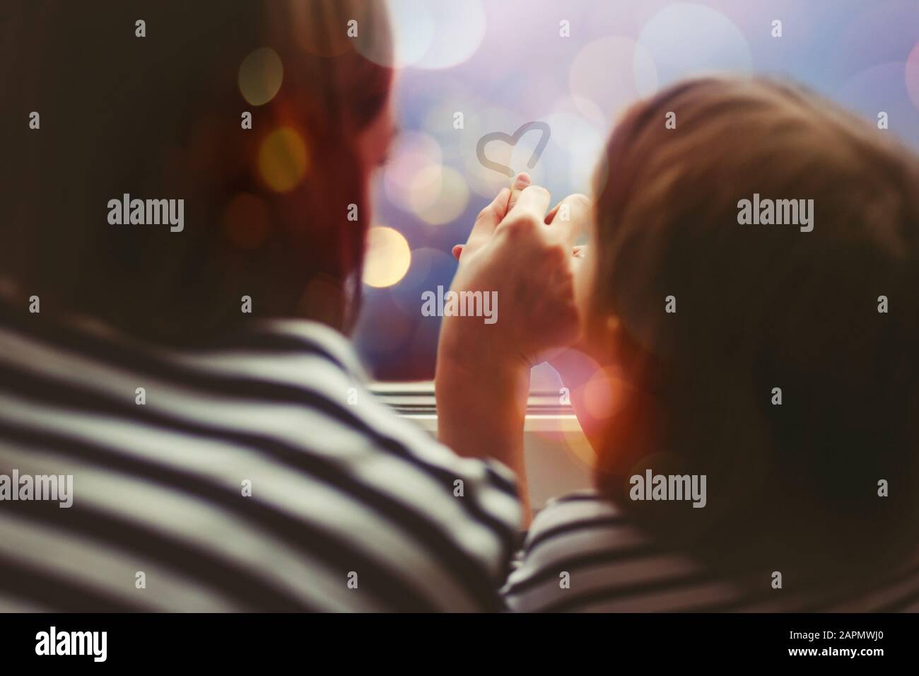 Little girl with her mother drawing heart symbol by fingers on misted window glass with evening lights behind glass. Selective focus. Stock Photo