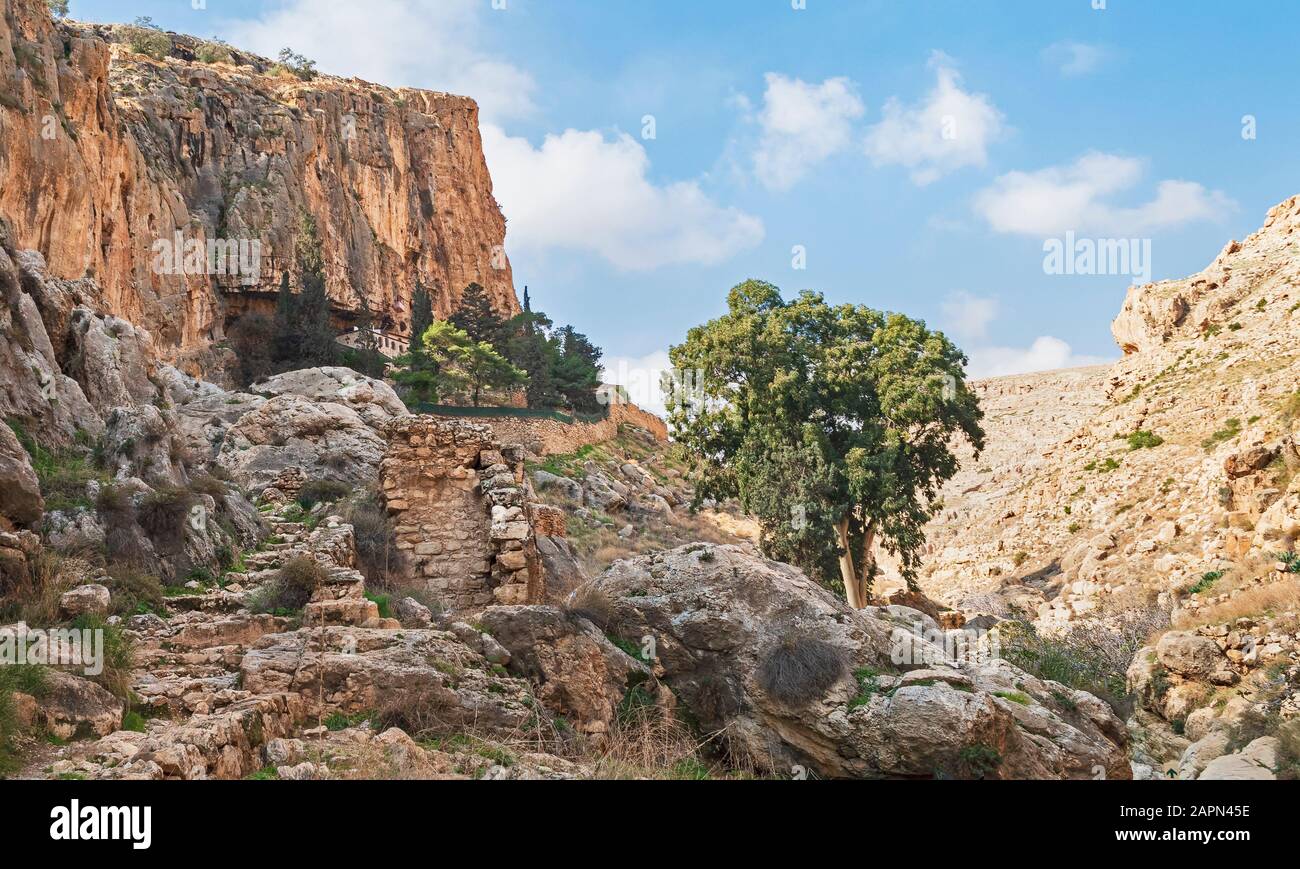 an ancient monastery clings to the side of a cliff near ein prat in wadi qelt in the west bank with an old stone wall in the foreground Stock Photo