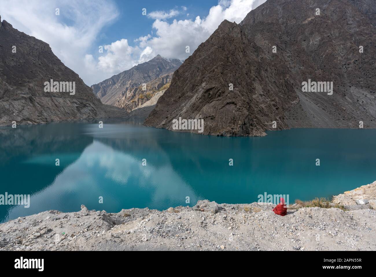 Pakistani woman in red hijab sitting on rocks by turquoise water of Attabad Lake in Gilgit Baltistan, Pakistan Stock Photo
