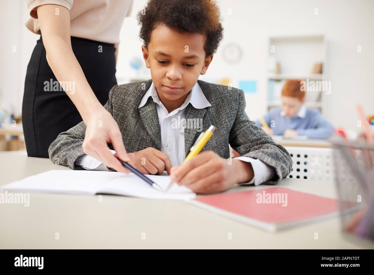 African schoolboy sitting at desk and writing in notebook with teacher explaining the material to him at the lesson Stock Photo