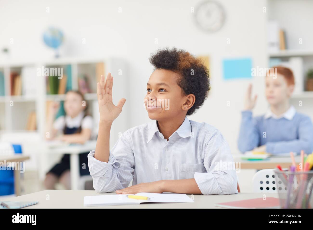 African smiling schoolboy sitting at his desk and raising his hand he ...