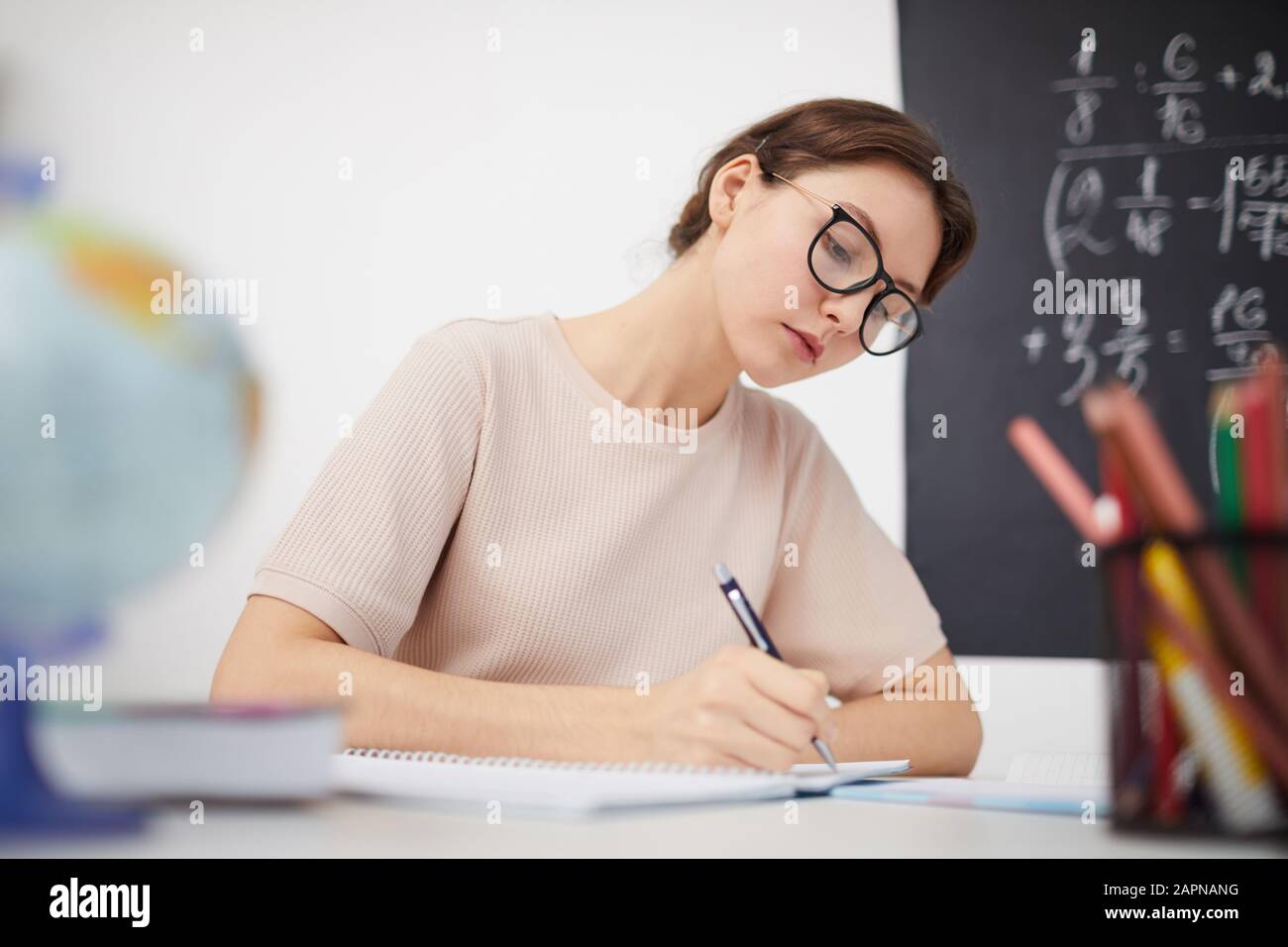 Young female teacher in eyeglasses sitting at the table and doing paper work in the classroom at school Stock Photo