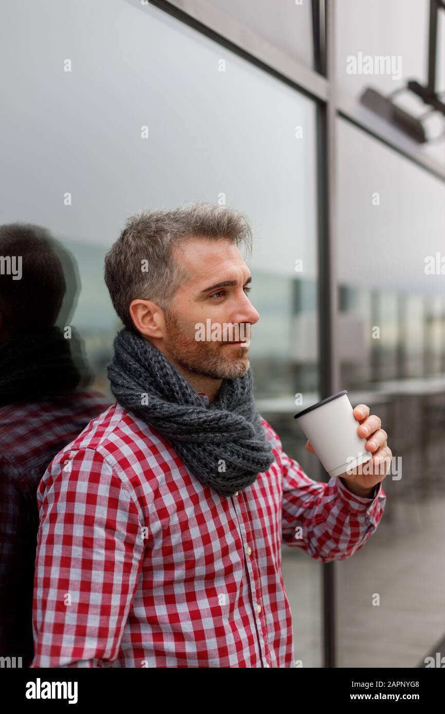 Young man leaning against a glass wall with a reusable glass in his hand Stock Photo