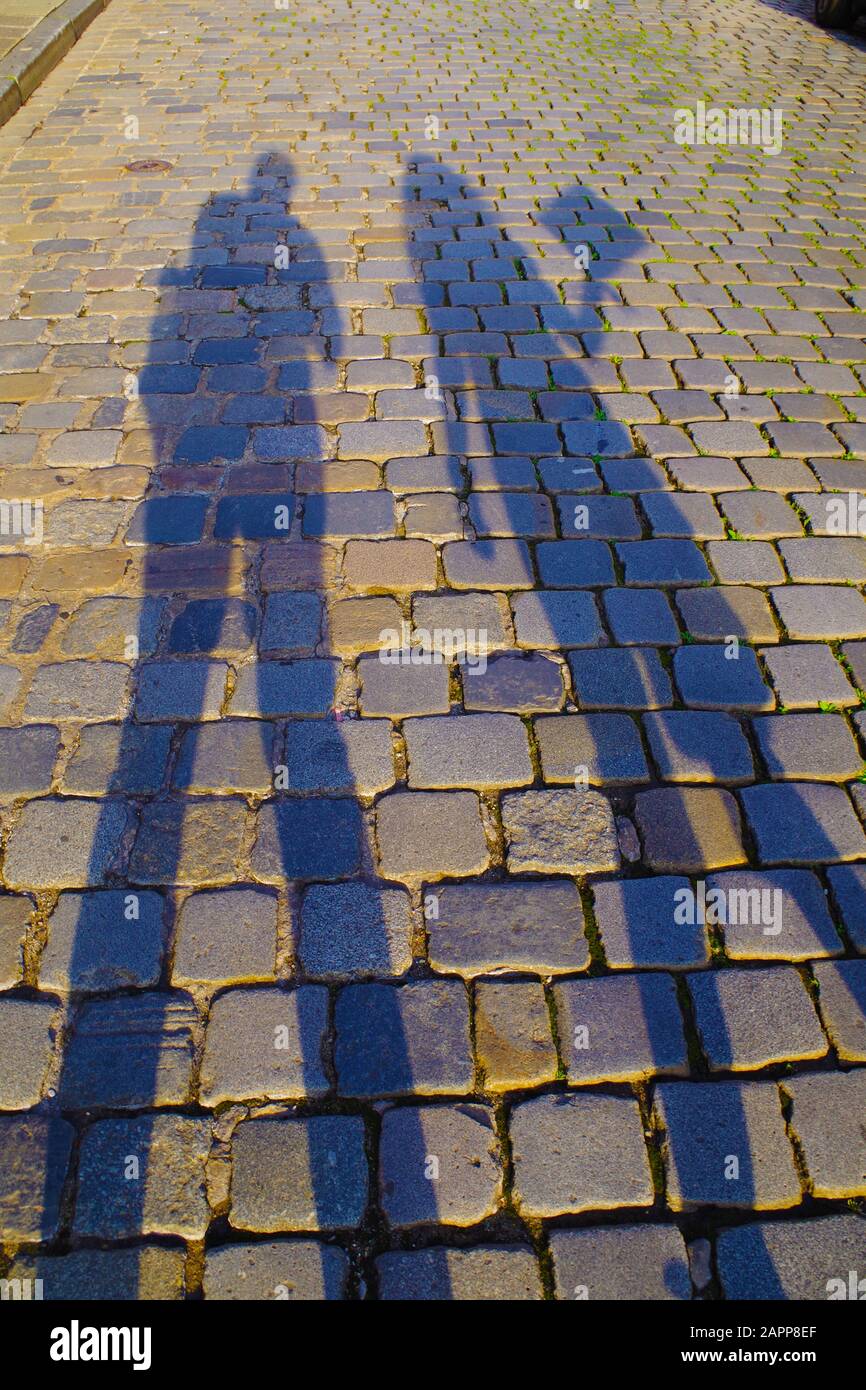 Dark, elongated shadows of man and woman on an old cobblestone pavement on a sunny day. Stones are colorful and reflect the sun rays. Stock Photo