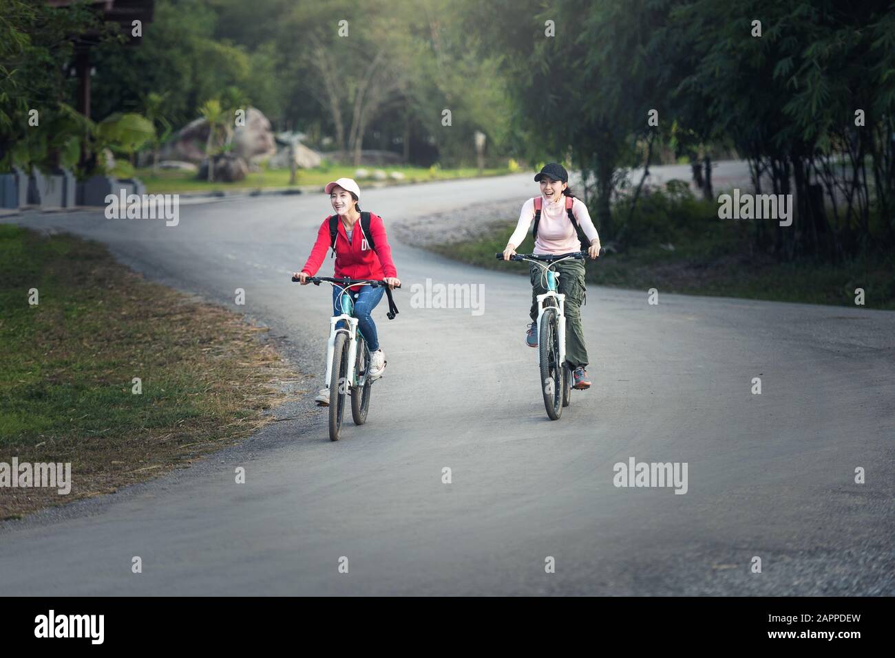 Two stylish young female friends on a bicycle along road. Best friends enjoying a day on bike. Stock Photo