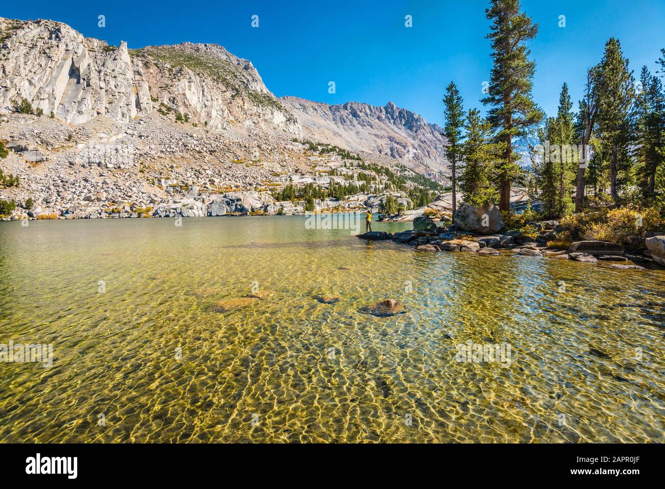 A hiker stands along the shore of Blue Lake, John Muir Wilderness, CA. Shear mountain peaks surround the green-blue lake. Stock Photo