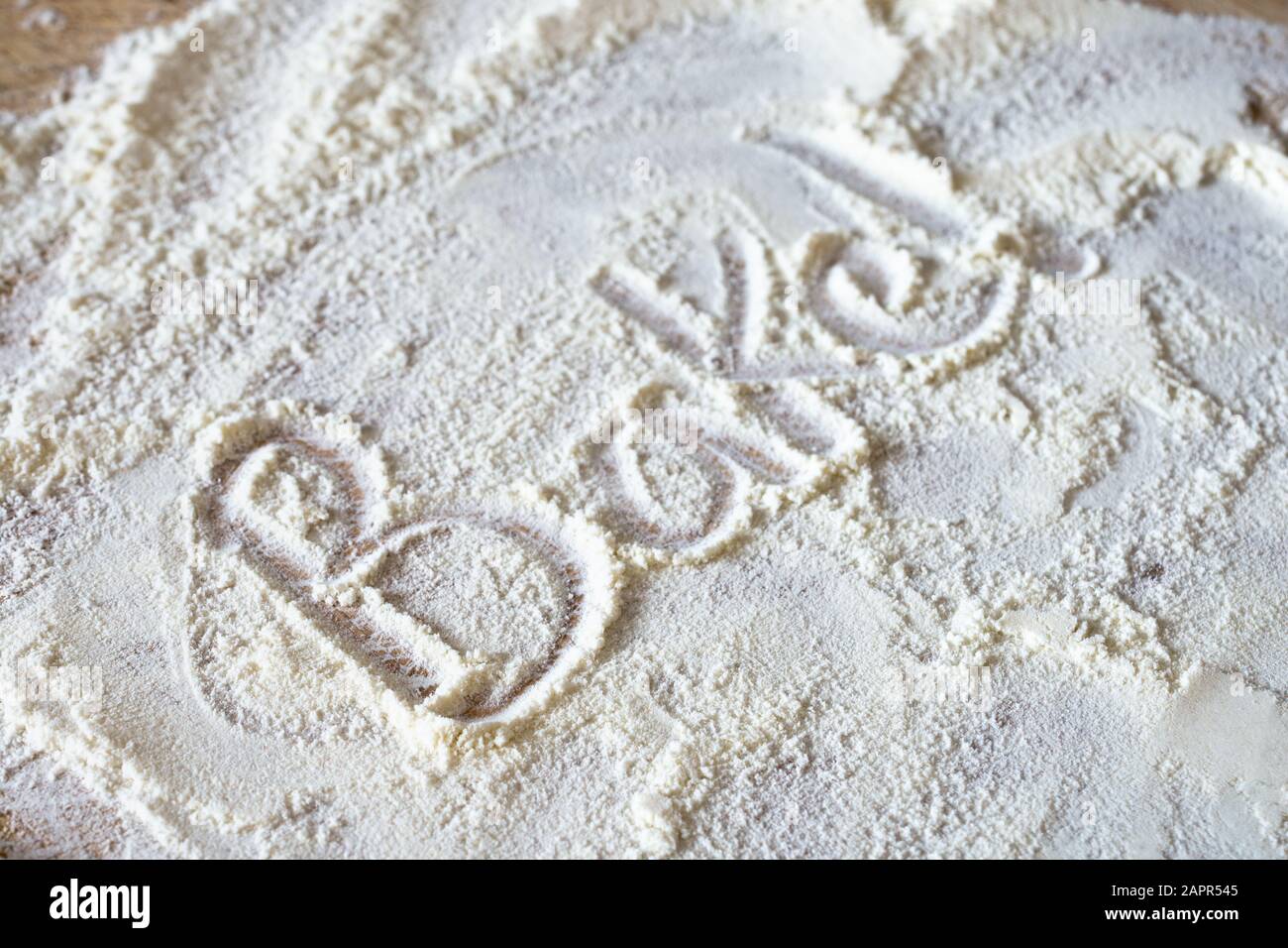 The word 'Bake!' written into messily scattered flour revealing wooden board below. Stock Photo
