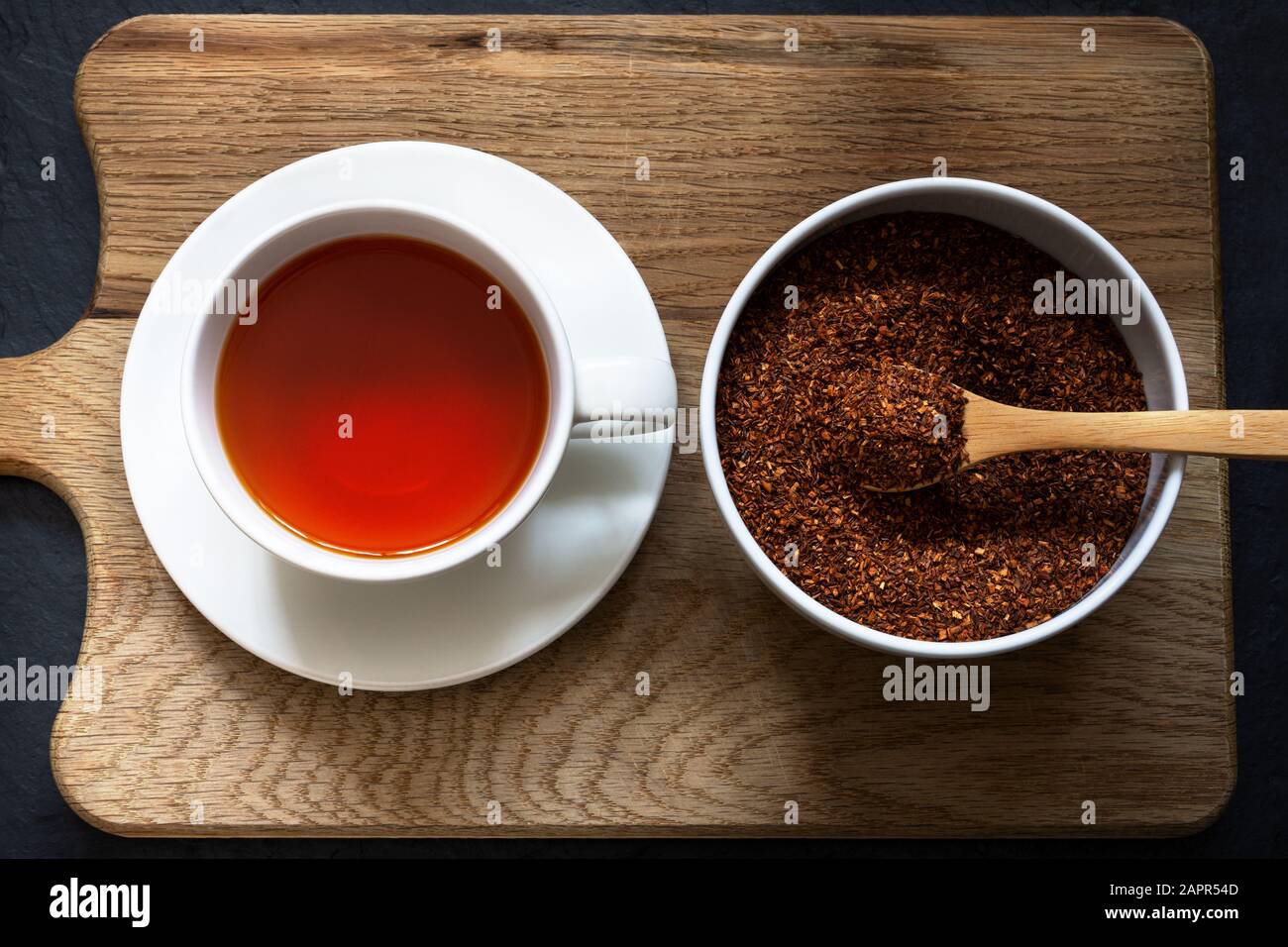 Overhead shot of a cup and saucer containing rooibos (redbush) tea with white bowl of leaves scooped in a wooden spoon. Wooden board with black slate Stock Photo