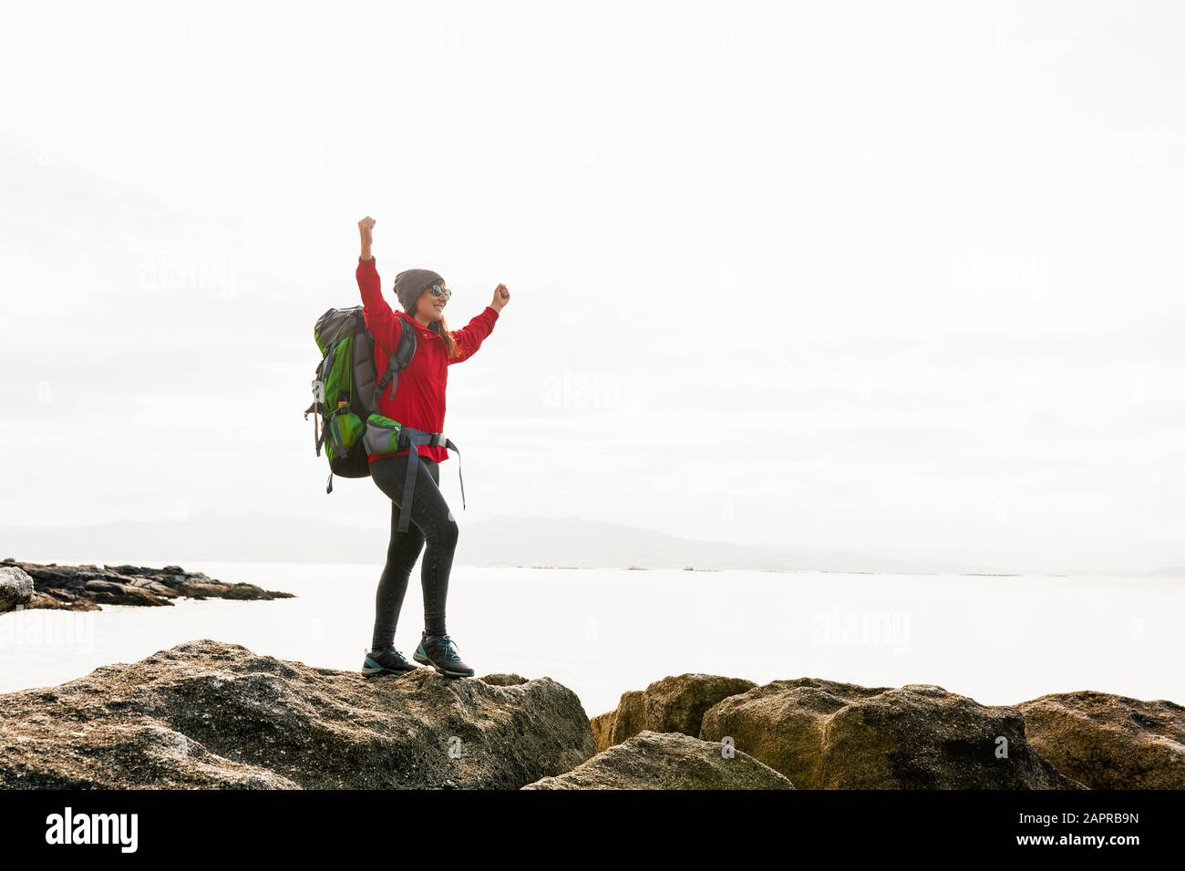 Woman with backpack and arms raised  enjoying the beautiful morning view of the coast Stock Photo