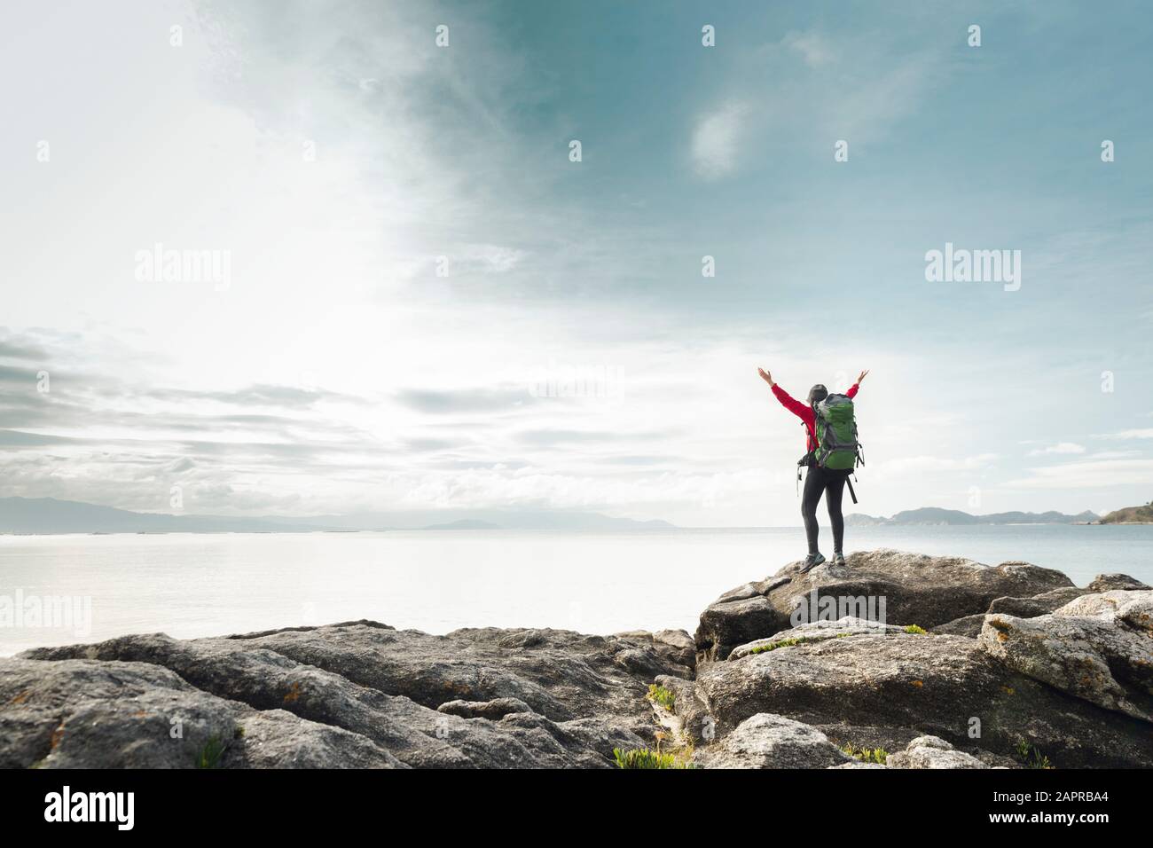 Woman with backpack and arms raised  enjoying the beautiful view of the coast Stock Photo