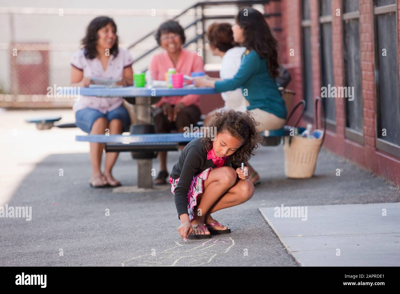 A girl crouches on the sidewalk and draws hopscotch game with chalk with three women sitting and talking behind her Stock Photo