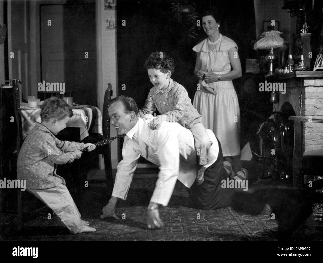 Domestic scene: Dutch family in the 1950s. Father plays in front of the coal stove with the children (riding horse”), while mother is smiling, [1956]. Stock Photo