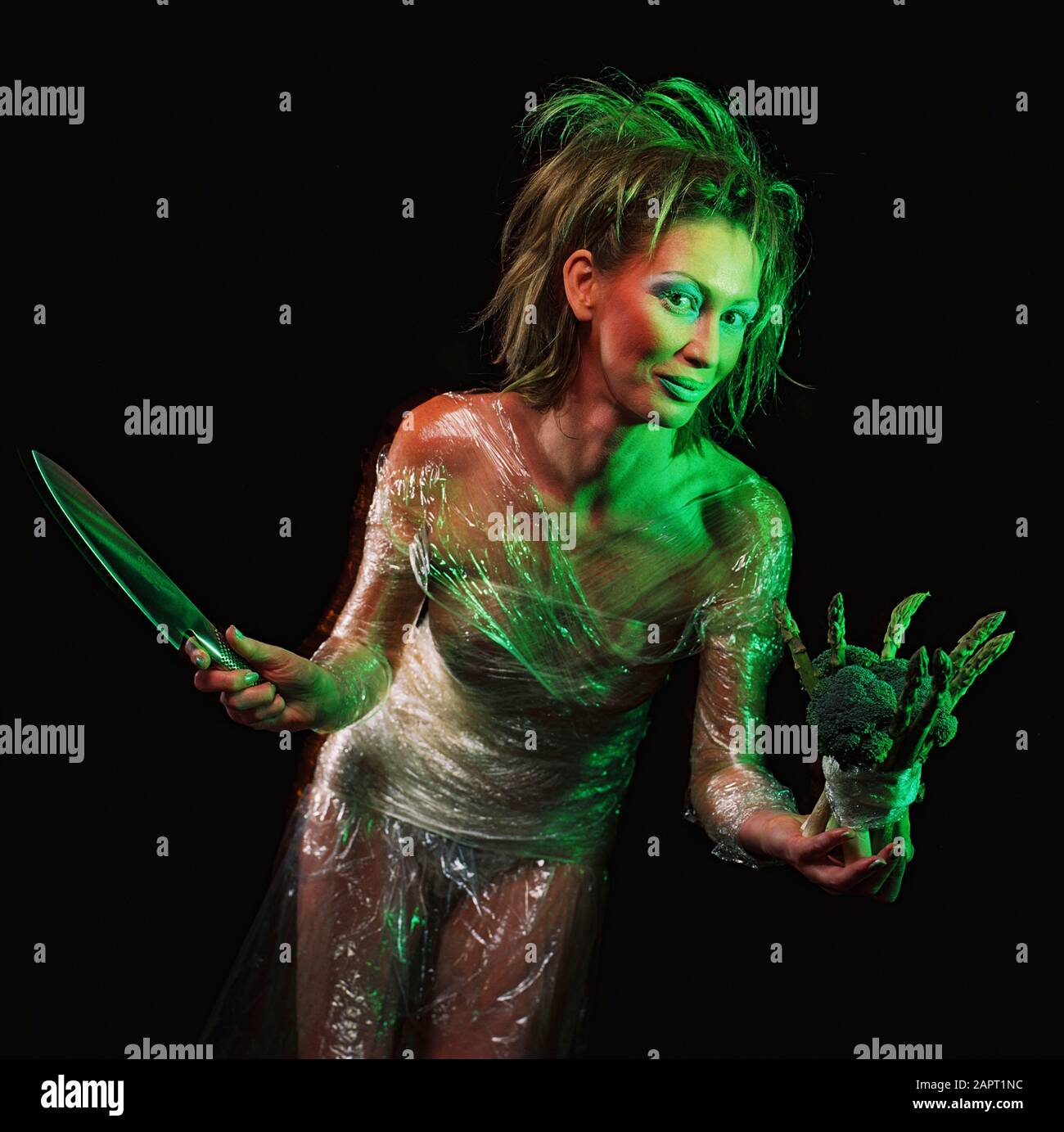 Studio image of young woman wrapped in plastic film and holding a knife a bunch of green vegetables. Stock Photo