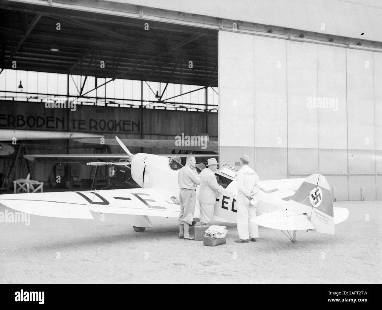 National Aviation School Holland  German Fieseler Fi5R low decker (D-EDIL) at Rotterdam Airport Waalhaven. The pilots are in conversation with the man in 'Standard' overall Annotation: In the hangar the inscription Forbidden to smoke Date: 1935 Location: Rotterdam, Waalhaven Keywords: architecture, hangars, pilots, smoking, airplanes, airports Stock Photo