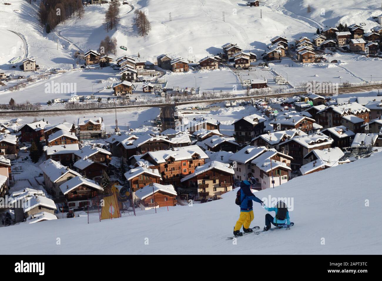 Two snowboarders on snowy ski slope in high winter mountains and sunlit village at background. Evening in Italian Alps. Livigno, region of Lombardy, I Stock Photo