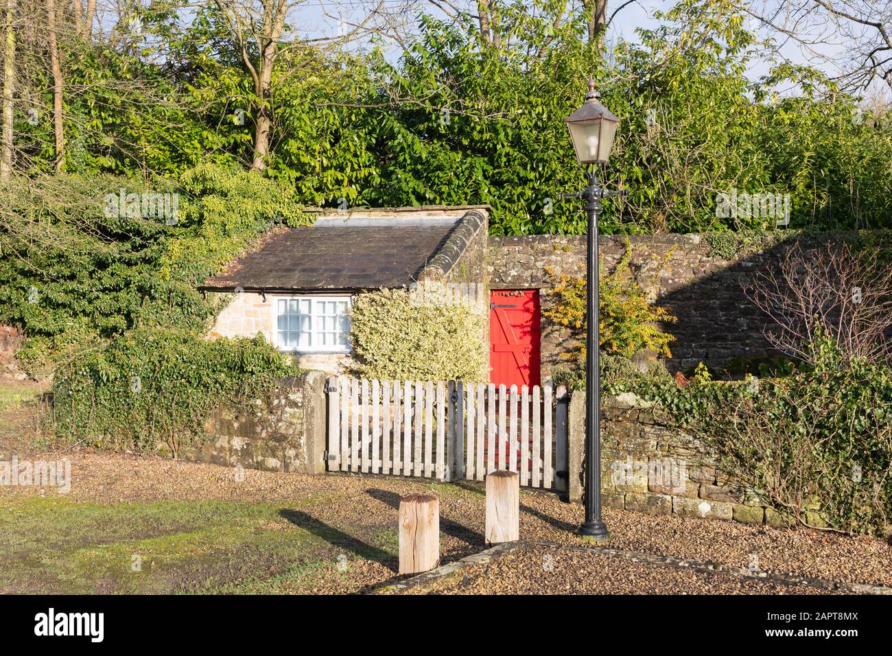 Cromford, Derbyshire, UK: Small, stone outhouse stands in a garden behind white gates and a stone wall. A lantern style streetlamp stands in front. Stock Photo