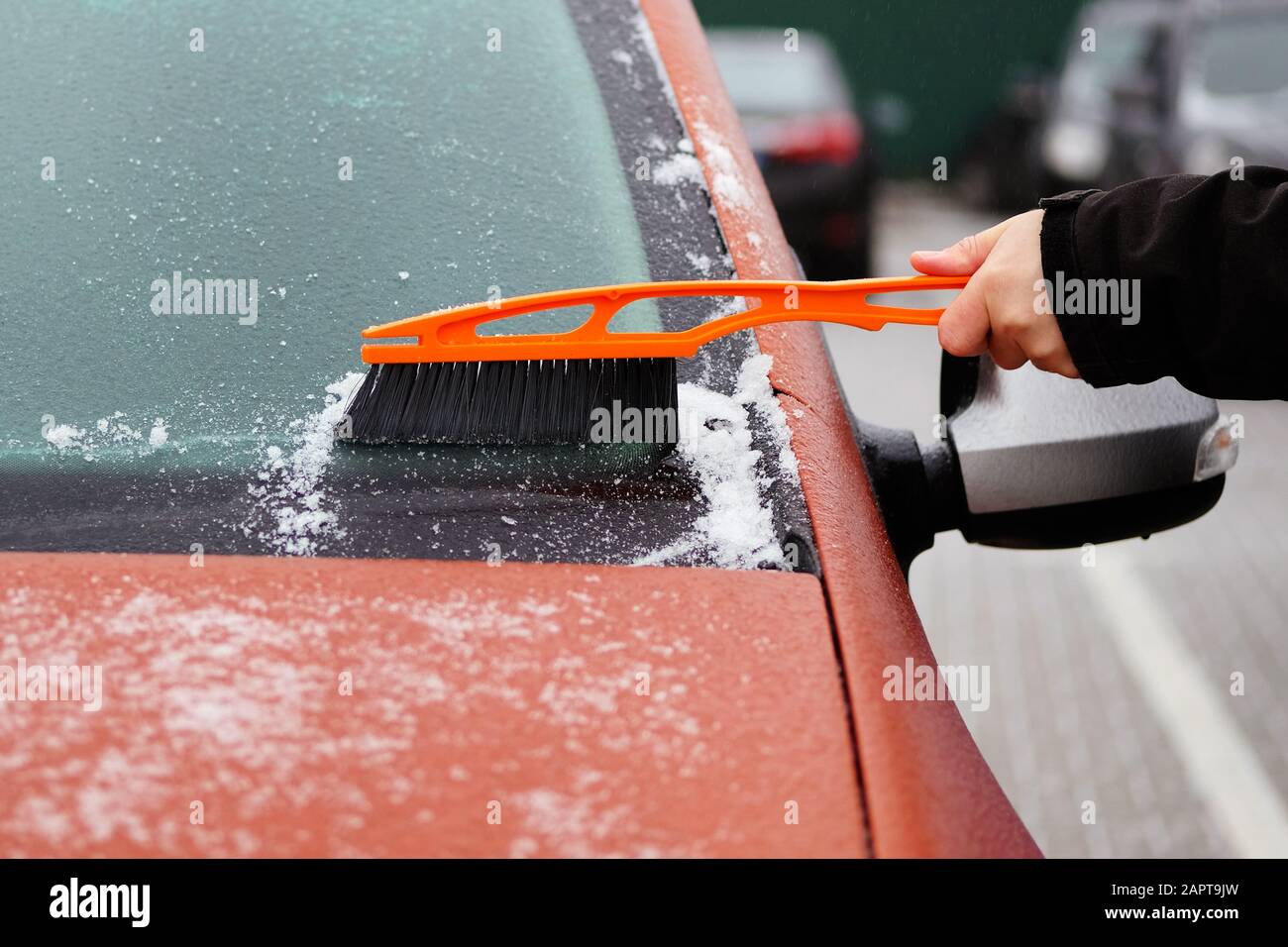 Man clears snow from icy windows of car. Orange brush in mans hand. Windshield of car, horizontal view. Stock Photo