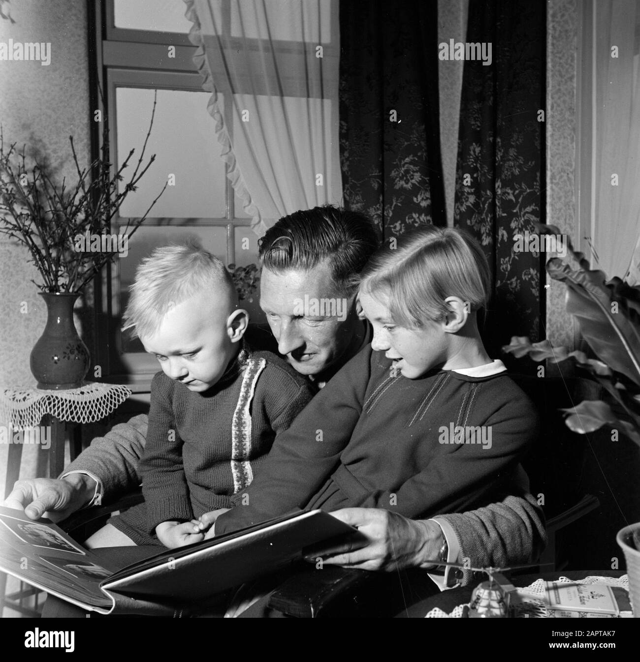 Gerard with his two children Gerrie and Marietje on lap while viewing a photo album Annotation: Gerard de Jong survived only a fusillade on April 7, 1945 at Dronrijp Date: 1 January 1955 Location: Friesland, Leeuwarden Keywords: pottery, books, interiors, children, tables Personal name: Young, Gerard de, Jong, Gerrie de Stock Photo