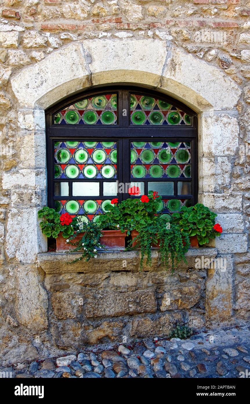 colorful glass window, arched, old stone building, windowsill, potted flowers, contrasting textures, Perouges, France, summer, vertical Stock Photo