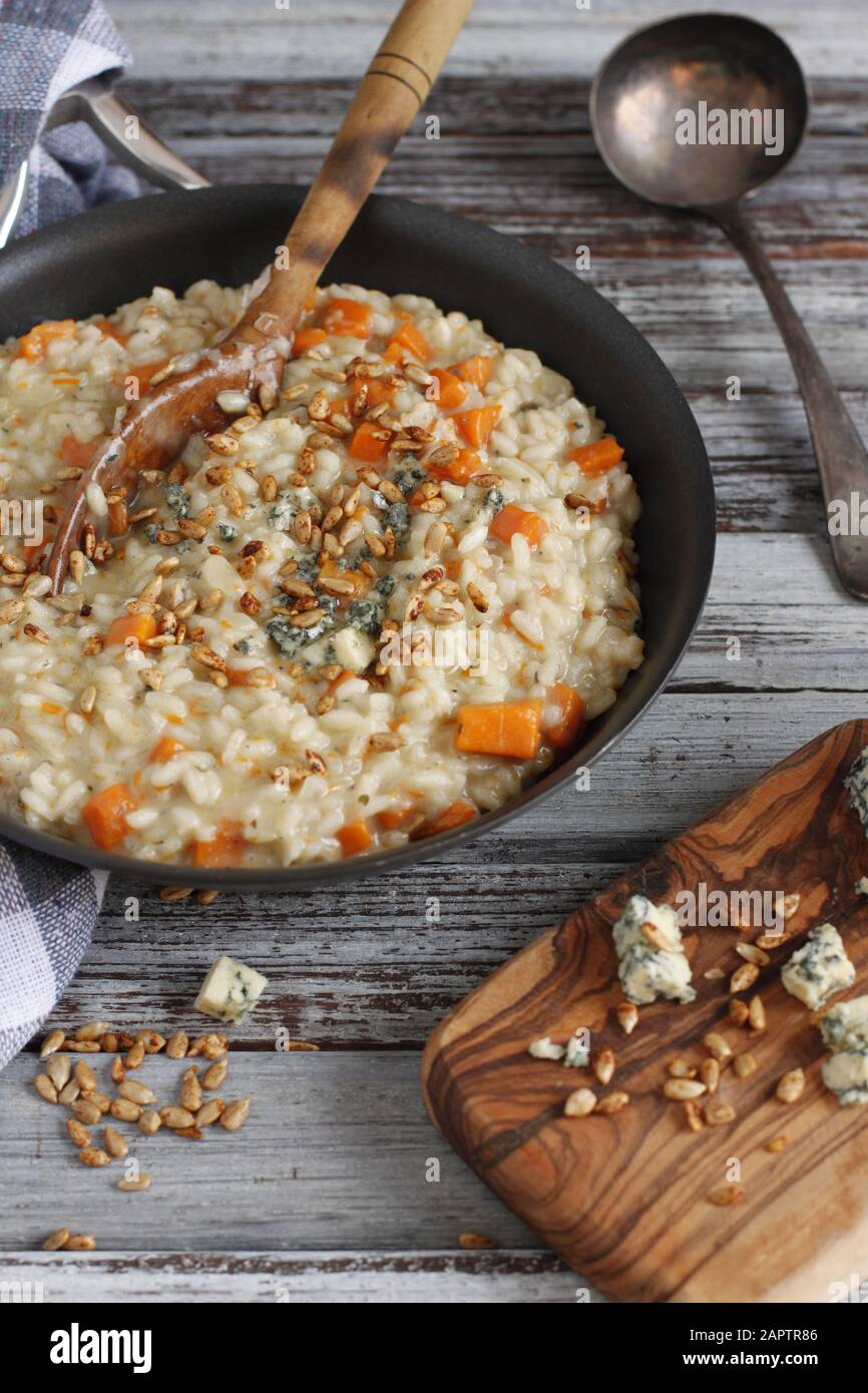 Butternut squash risotto in pan with wood spoon on rustic background with board, cheese, sunflower seeds and ladle Stock Photo