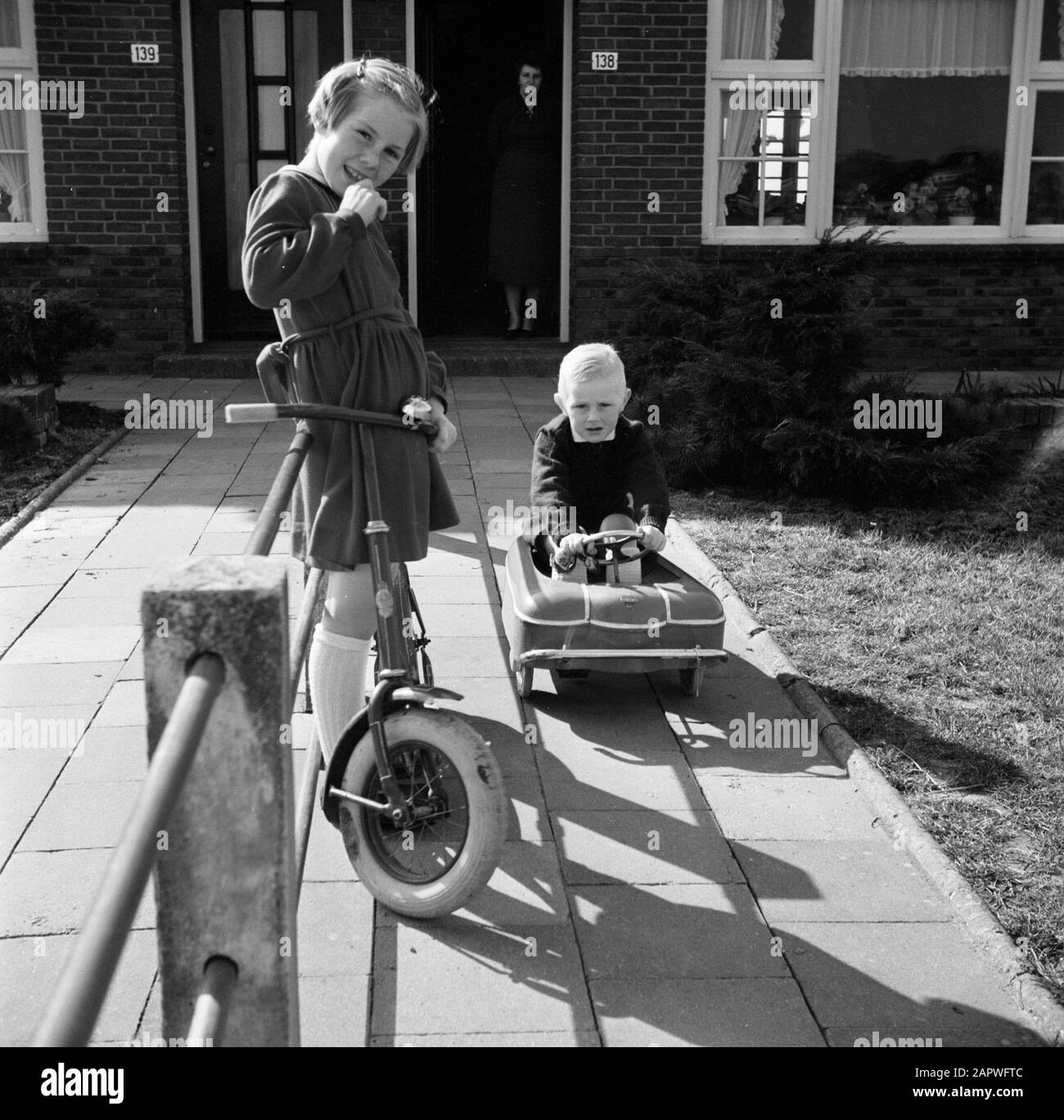 Marietje, the eight-year-old daughter of Gerard, on her autoped in the front garden, together with her four-year-old brother Gerrie. Her mother watches in the doorway Annotation: Gerard de Jong survived a fusillade on 7 April 1945 at Dronrijp Date: 1 January 1955 Location: Friesland, Leeuwarden Keywords: autopeds, doors, children, women, homes Personal name: Jong, Gerrie de, Jong, Marietje de, Jong,  the Stock Photo