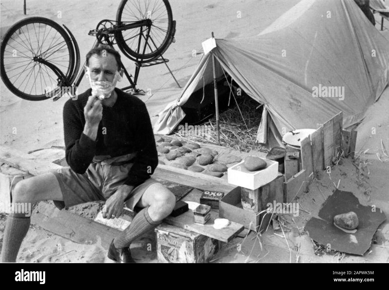 A camper shaves at his tent. In the background there is a bike upside down in the sand. England, circa 1925-1935. Stock Photo