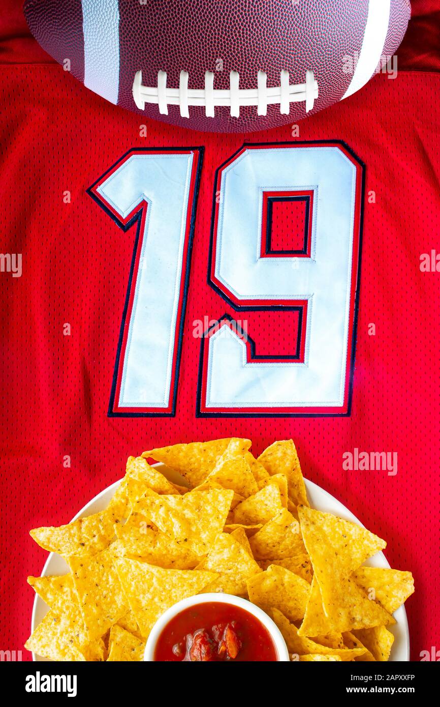 An American football with organic nacho chips and mild salsa on a white red football jersey with the 19 number Stock Photo