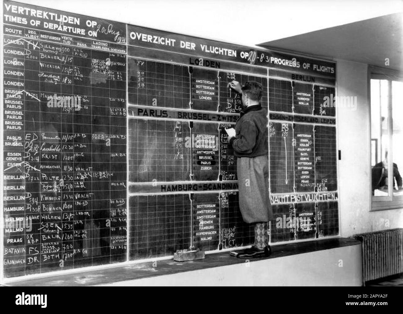 Schiphol for 1945. Information board for arrivals and departures of aircraft, in Schiphol station building. The photo shows young man dressed in a plusfour (puffed pants). He stands on a wide high edge along the wall. Against the wall is a large board (blackboard) where he keeps track of the times of the departing and incoming planes with white chalk. Schiphol 1931. Stock Photo