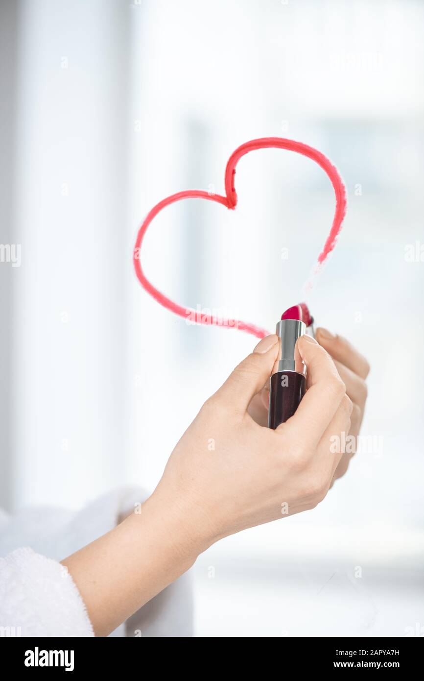Hand of flirty female with lipstick of crimson color drawing heart on mirror Stock Photo