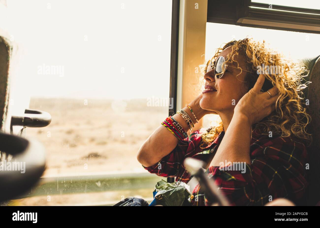 Beautiful blonde adult woman enjoying the travel on bus or train listening music with headphones - wanderlust lifestyle and happy people doing trip - Stock Photo