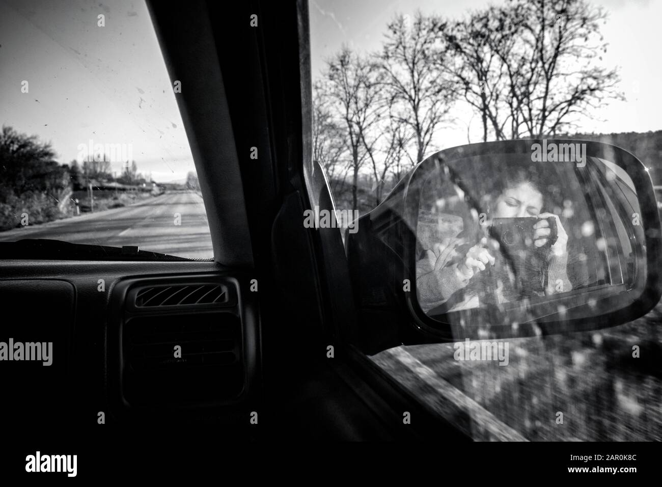 Female photographer taking a photo of herself in the right rearview mirror of a moving car driving in a conventional road. Black and white. B&W.Selfie Stock Photo