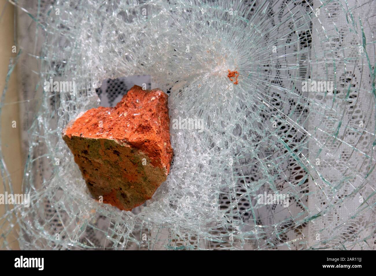 window with net curtains smashed with old style red brick in an act of vandalism hate crime criminal damage Stock Photo