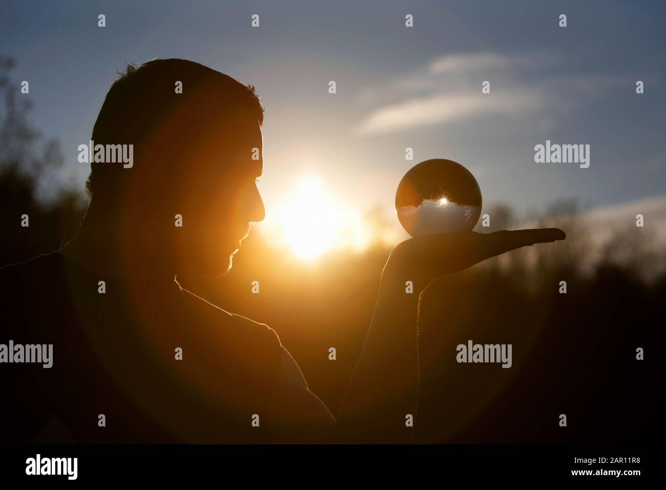 man holding and looking into glass orb as the sun sets Stock Photo