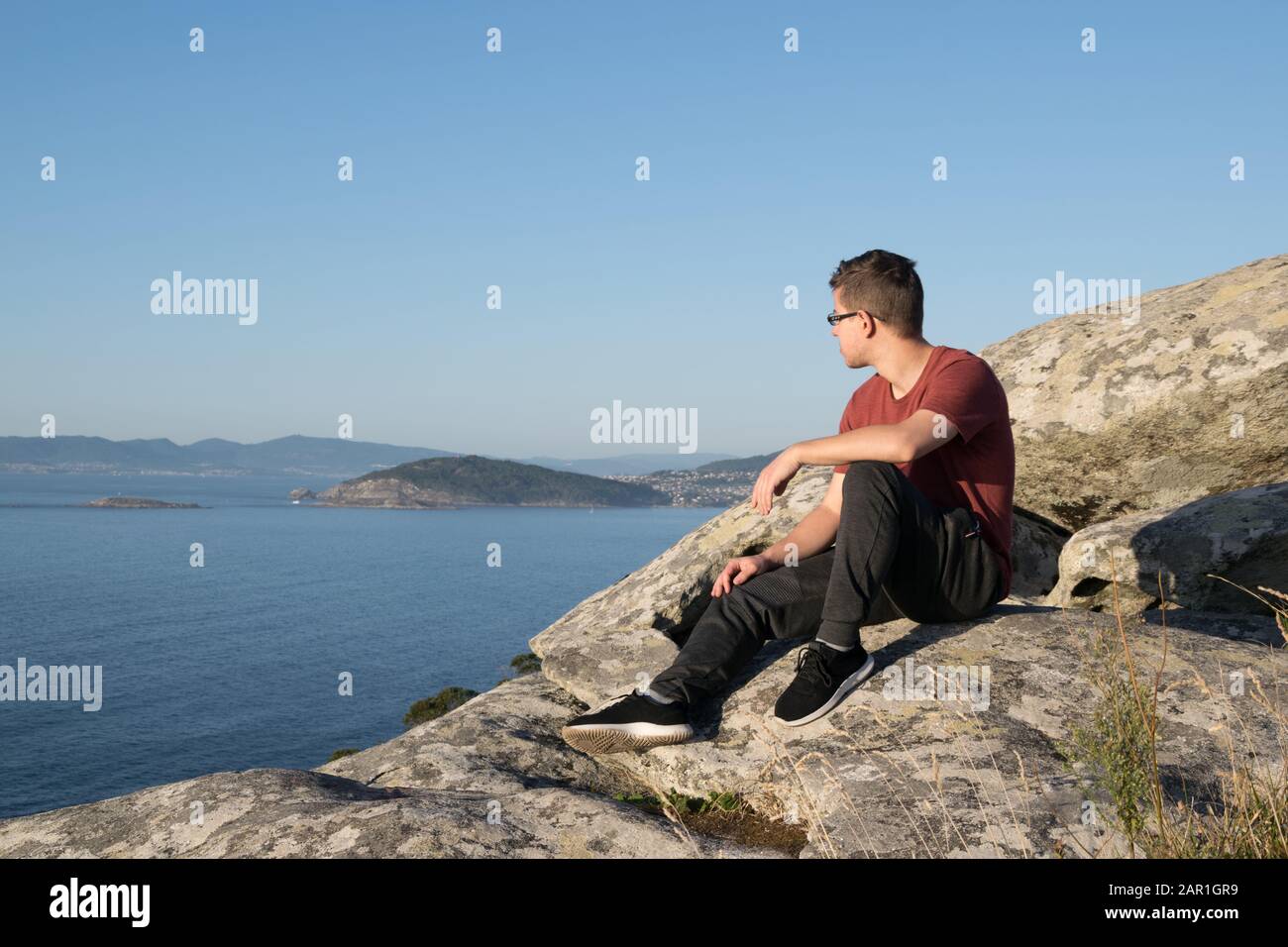 Boy sitting on the rocks of a mountain looking at the ocean Stock Photo ...