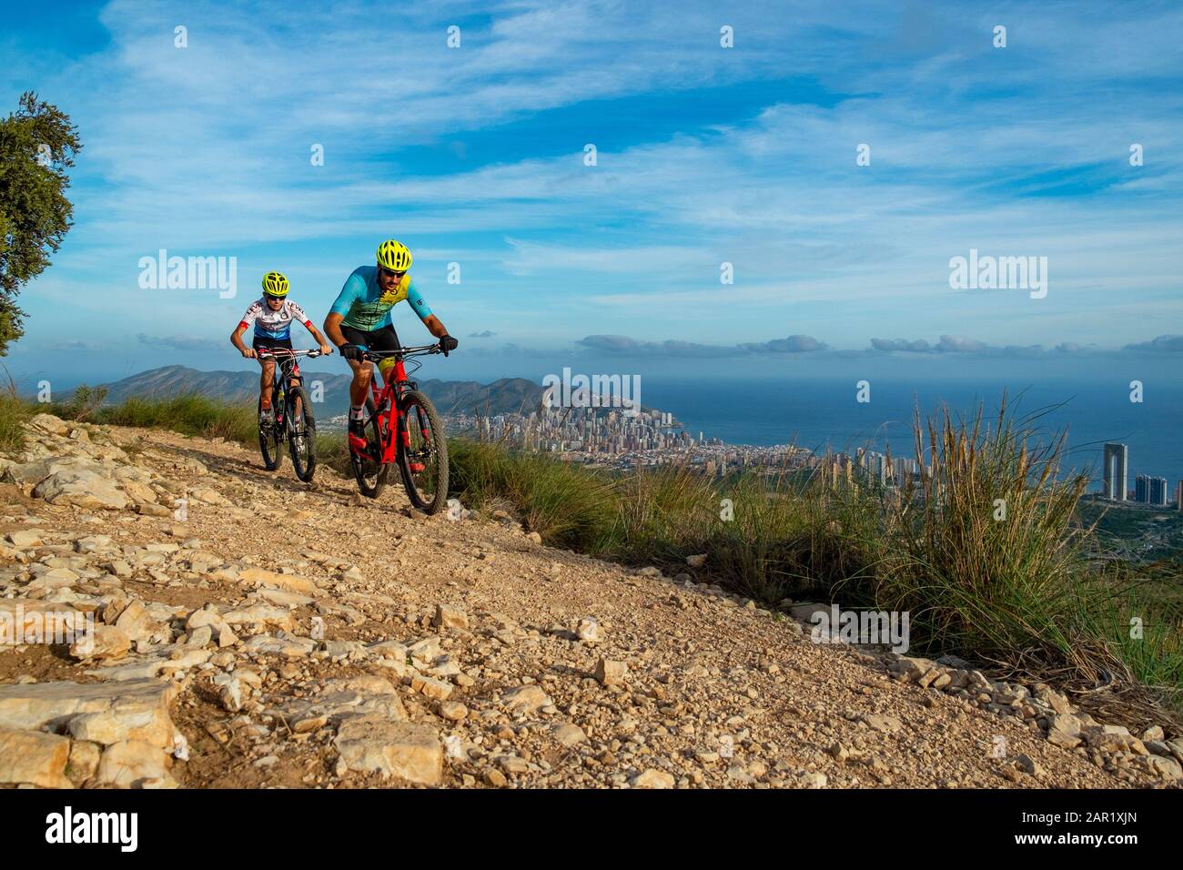 Two mountain bikers riding on hill with Benidorm city in the background, Sierra Cortina, Benidorm, Alicante province, Spain Stock Photo