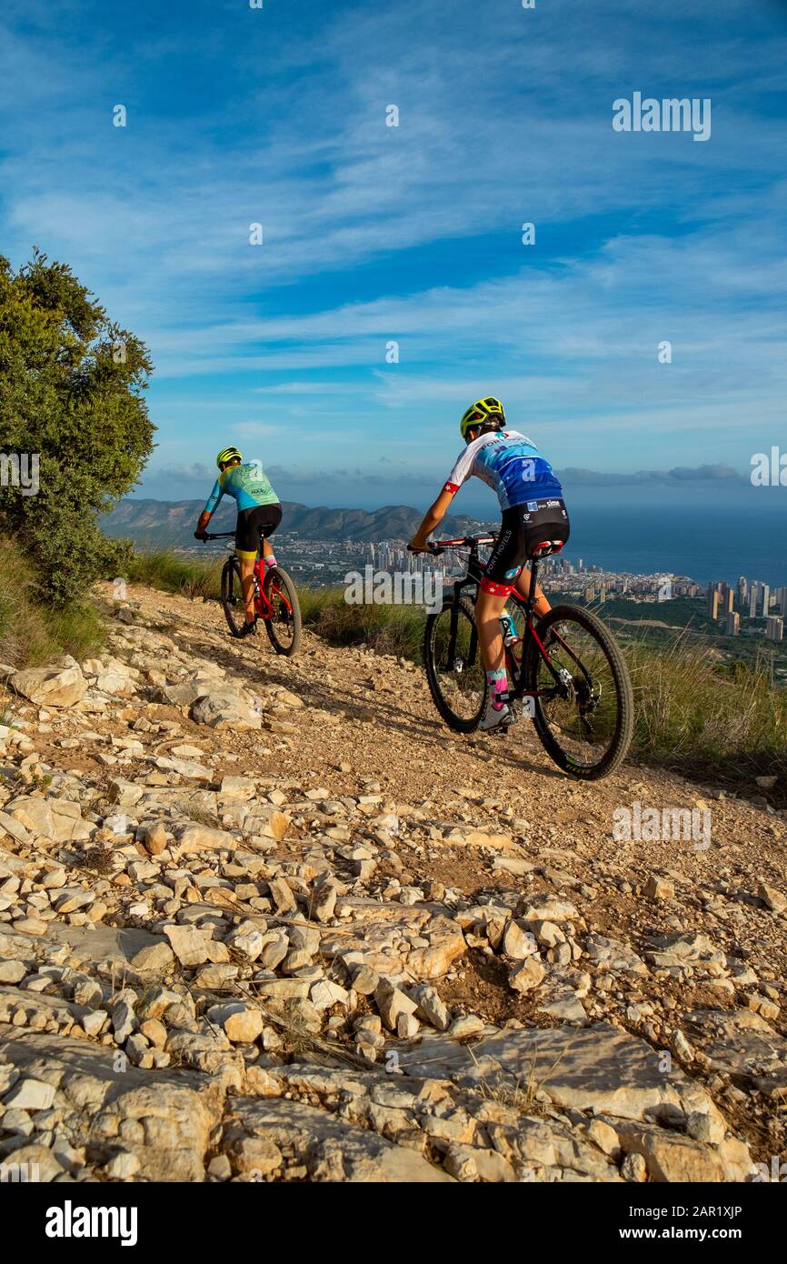 Two mountain bikers riding on hill with Benidorm city in the background, Sierra Cortina, Benidorm, Alicante province, Spain Stock Photo