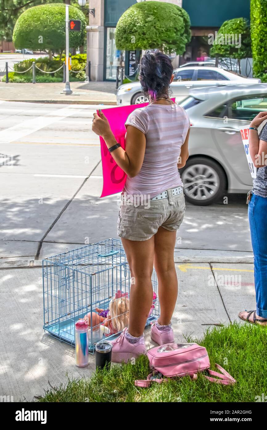 Girl dressed in pink and shorts stands on urban street corner with protest sign and a dog cage with dolls inside at rally against immigrant children i Stock Photo
