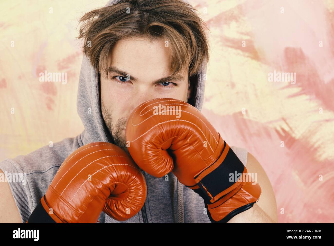 Man with messy hair on colorful background. Sports, box and strength concept. Guy in grey sleeveless hoodie wears red leather boxing gloves. Boxer with serious face trains to hit Stock Photo