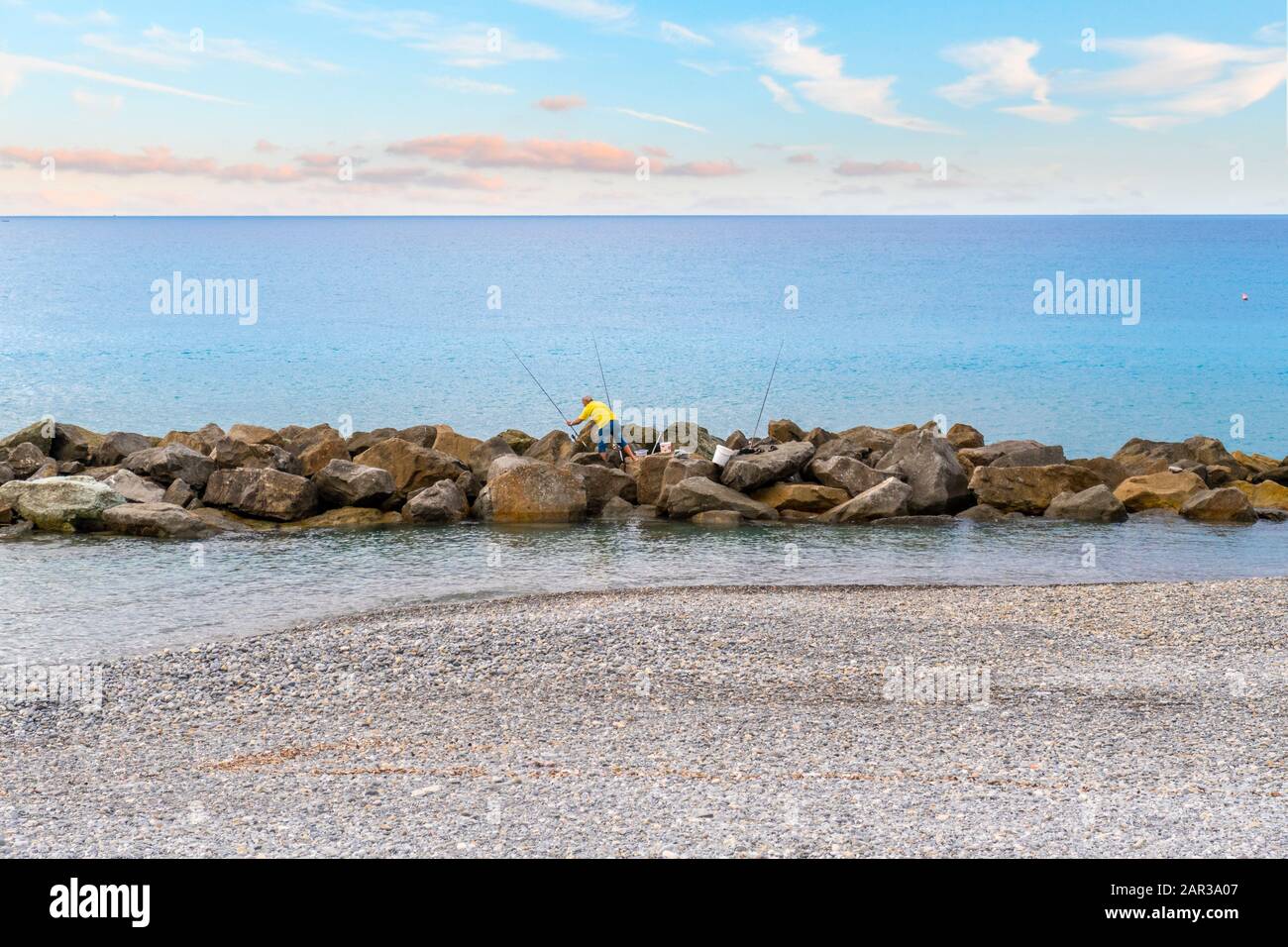 An Italian fisherman on the rocks at a pebble beach on the Mediterranean Sea at the town of Ventimiglia, Italy, on the Italian Riviera. Stock Photo