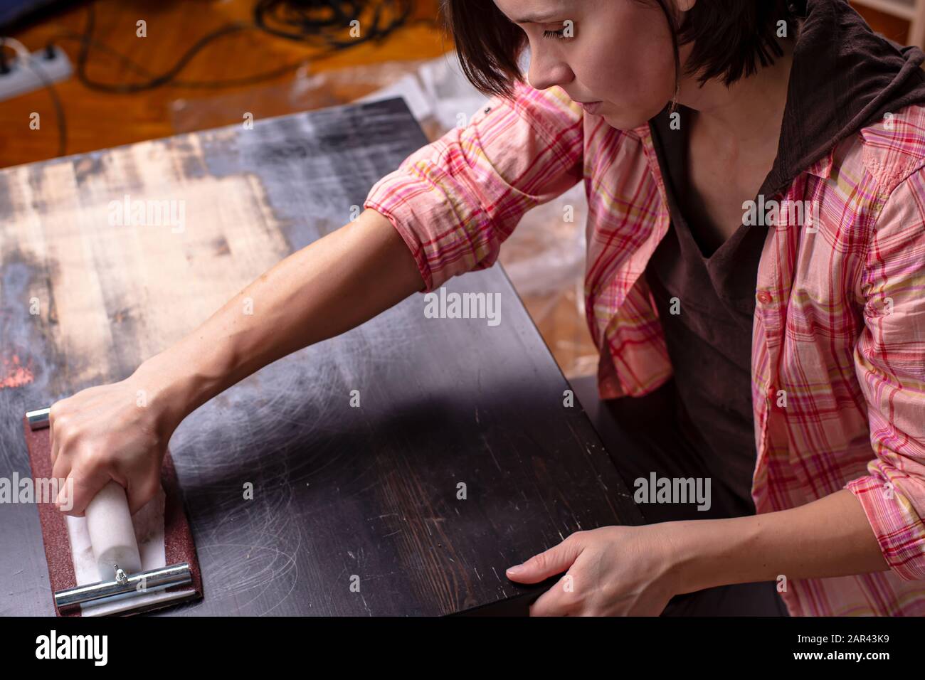 Woman prepares the surface of an old wooden black table for painting, using a carpentry holder for sandpaper. Home repair. Stock Photo