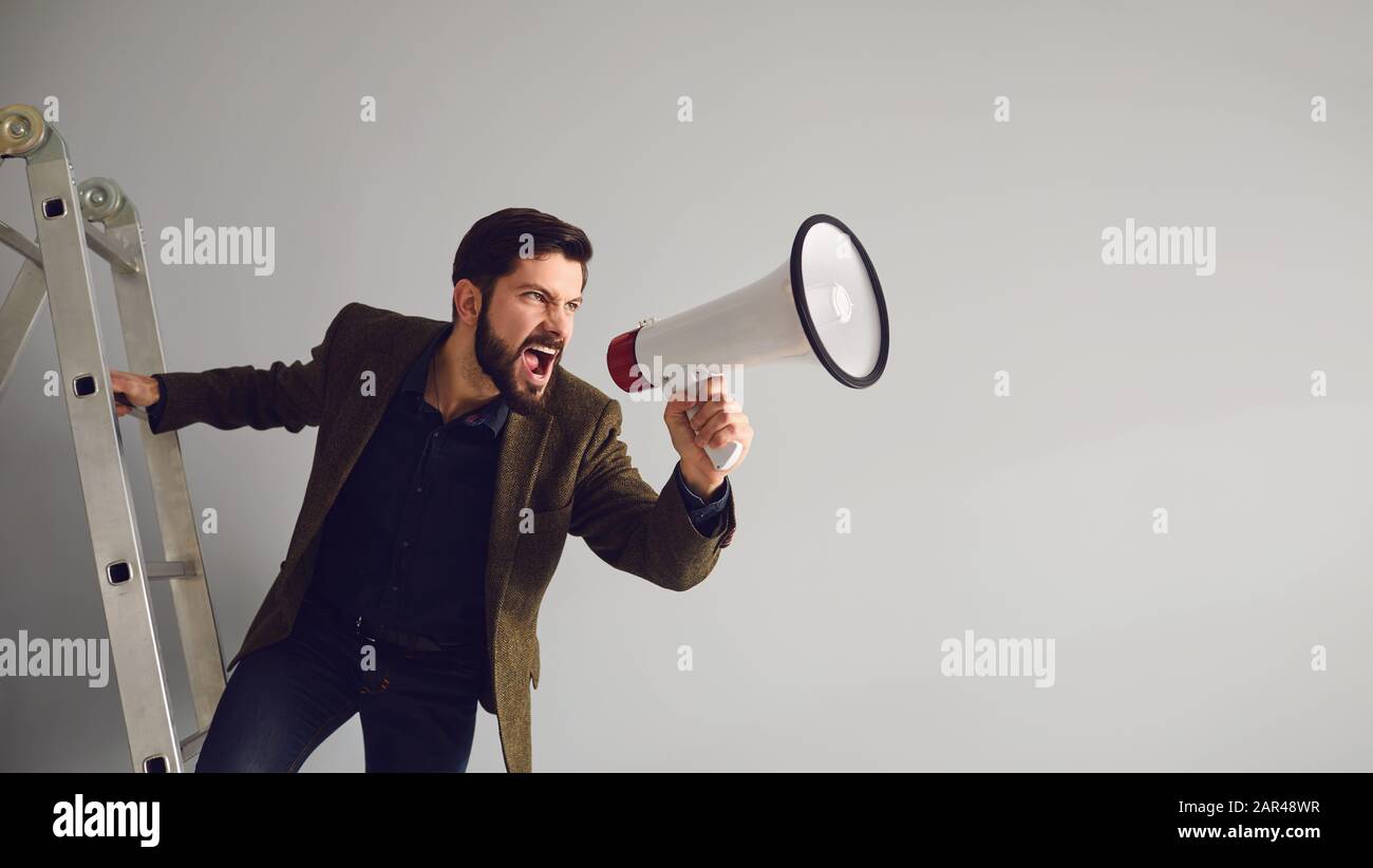 A bearded businessman on the stairs with a megaphone in hand shouts on a gray background. Stock Photo