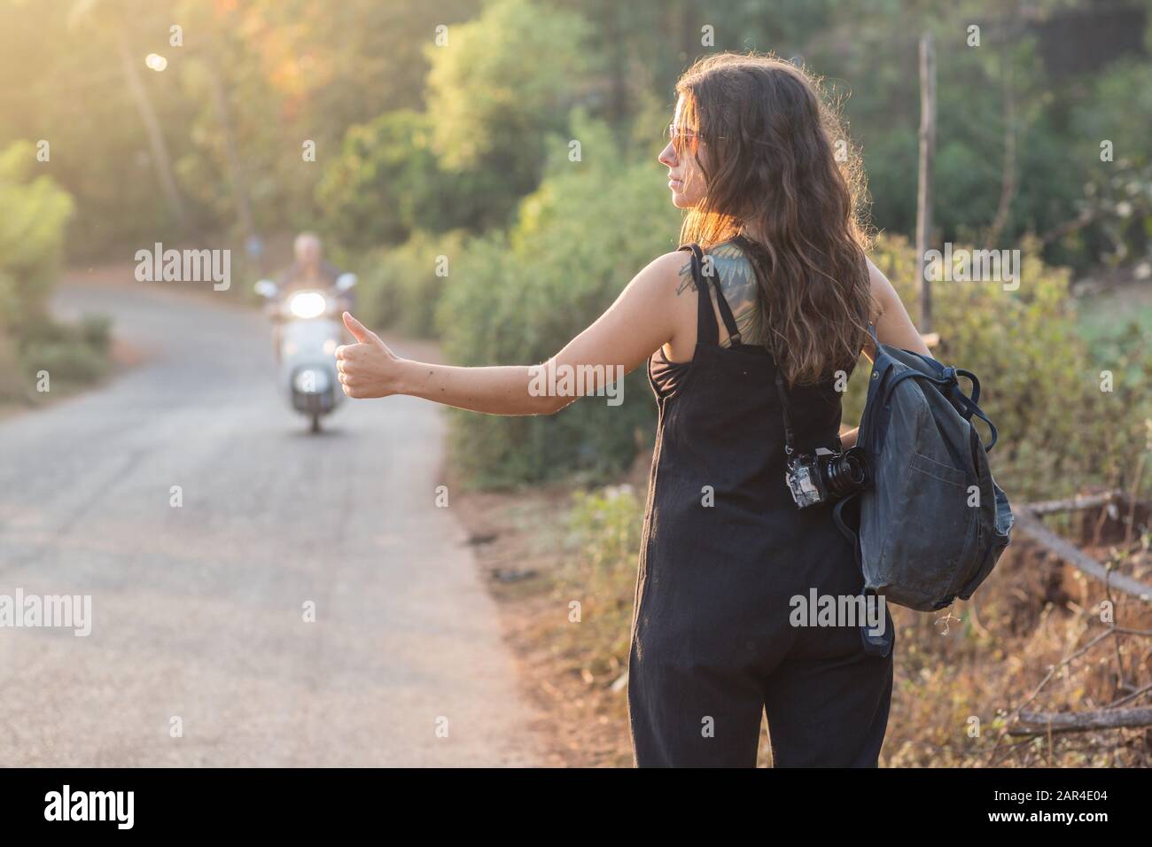 A girl with a backpack and camera hitchhiking in the sunset. Stock Photo